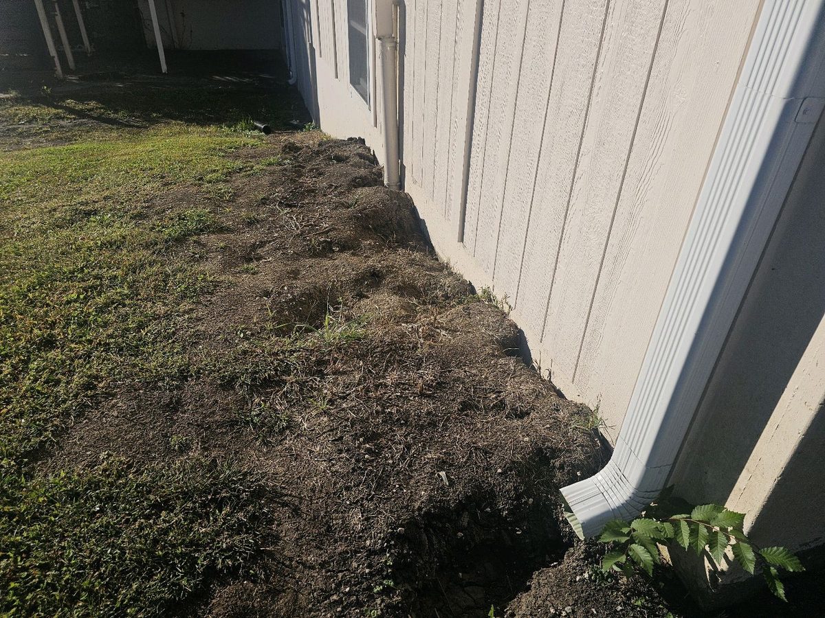 Mounded soil and patchy grass piled against a house's siding, with a downspout at the corner