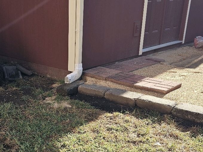 Downspout ending at the base of a wall with no extension, beside a brick patio edge, found during a home inspection in Olathe, KS